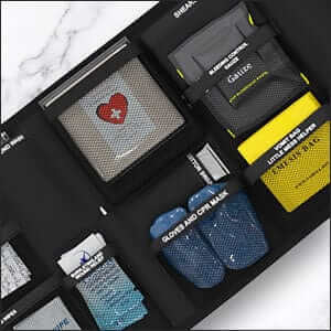 Family first aid kit including a Triangular bandage with the life safety pro heart-shaped design, gloves, and a pouch on a white marble surface. Also showing Vomex emesis bag, gloves, burn gel and hemostatic guaze. 